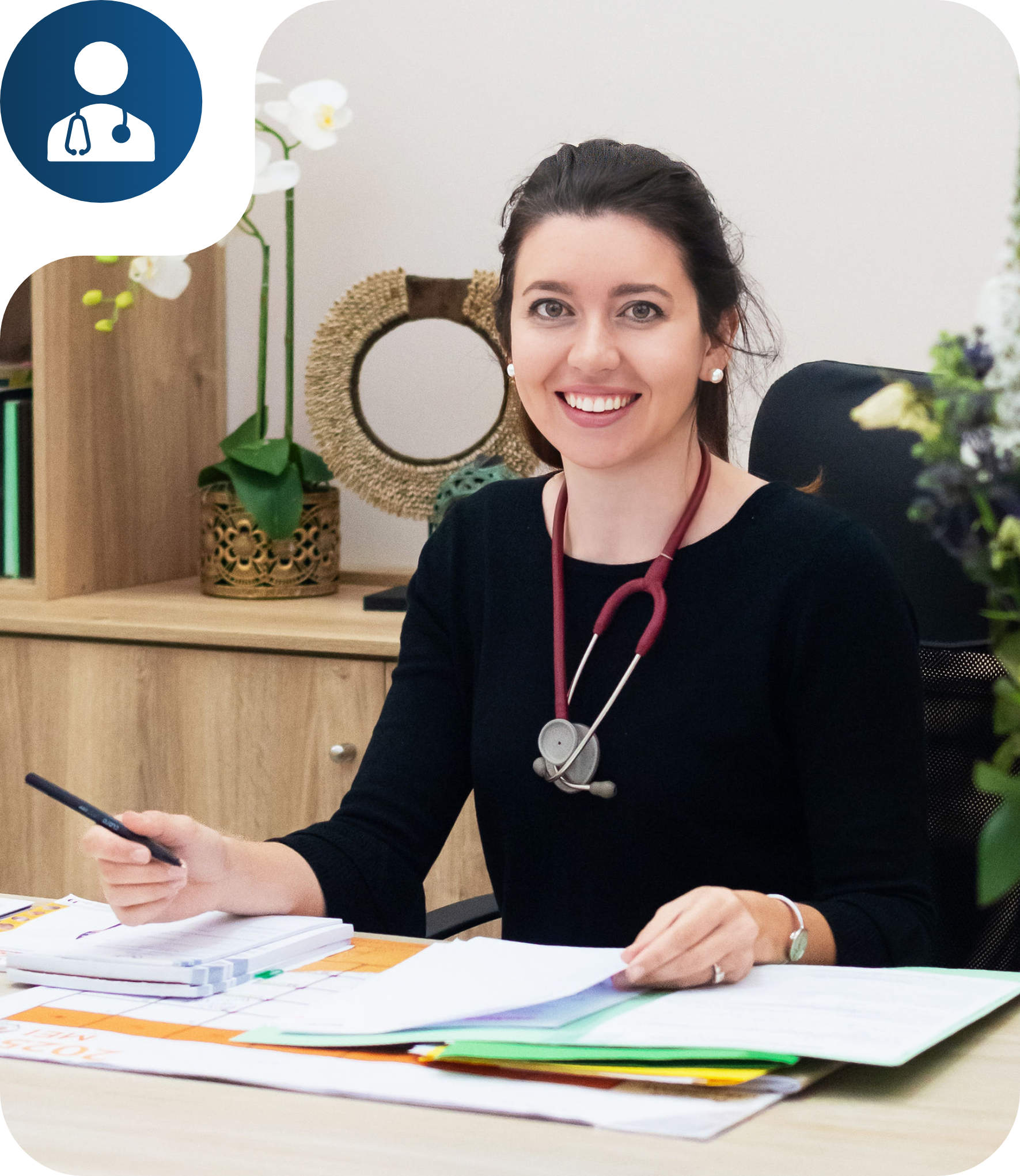 Smiling female doctor with a stethoscope around her neck sitting at a desk holding a pen and papers.